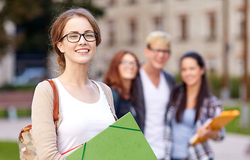 Eine jungen Frau mit Brille und einer Mappe in der Hand steht im Vordergrund. Sie lächelt. Auswahlthema: Voraussetzungend er Sozialassistentenausbildung.