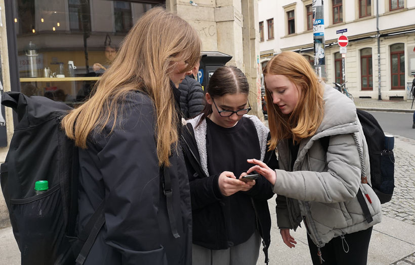 Drei angehende Sozialassistentinnen befinden sich auf einer Straße in der Dresdner Neustadt und beugen sich gemeinsam über das Display eines Mobiltelefons.